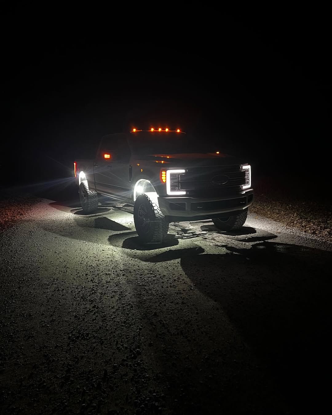 Pure White 9 LED rock lights glowing white in the wheel wells of a lifted Ford pickup on a dark gravel road at night