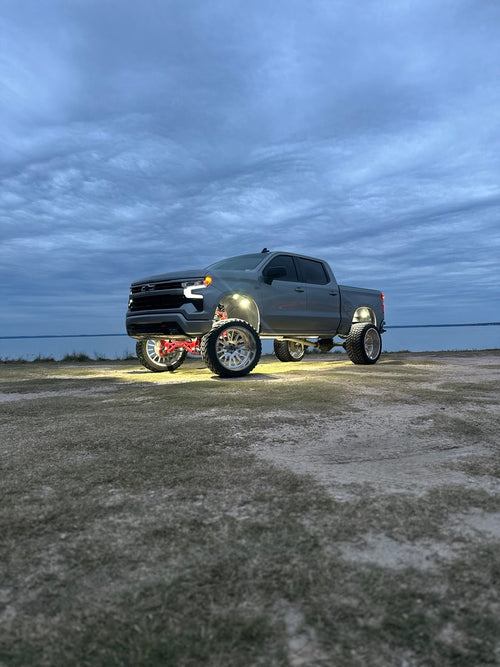 Pure White 36 LED rock lights glowing under a lifted Chevy pickup with red suspension parts, parked by the water at dusk