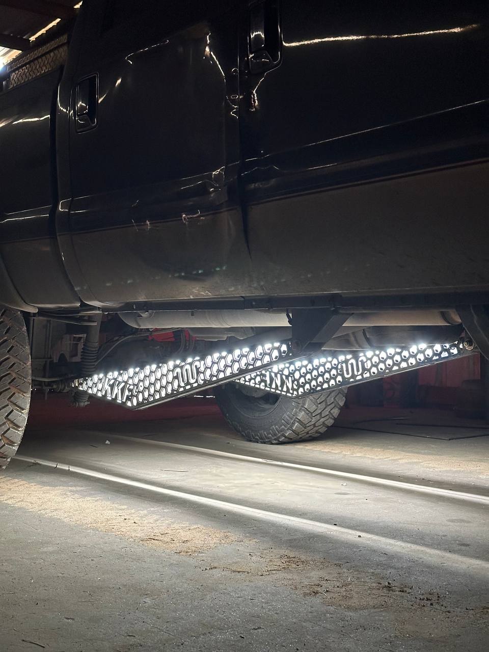 Switchback trac bar lights in white running light mode on both ladder bars of a black lifted pickup in a dark shop